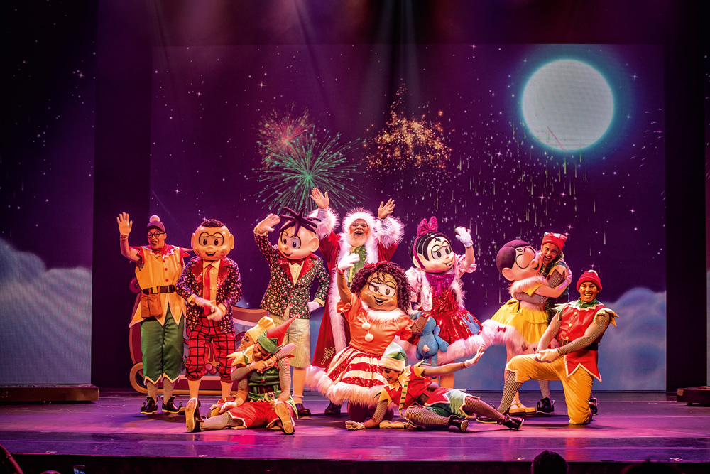 Em um palco de teatro, mais de dez pessoas estão vestidas de bonecos da Turma da Mônica e com enfeites de Natal