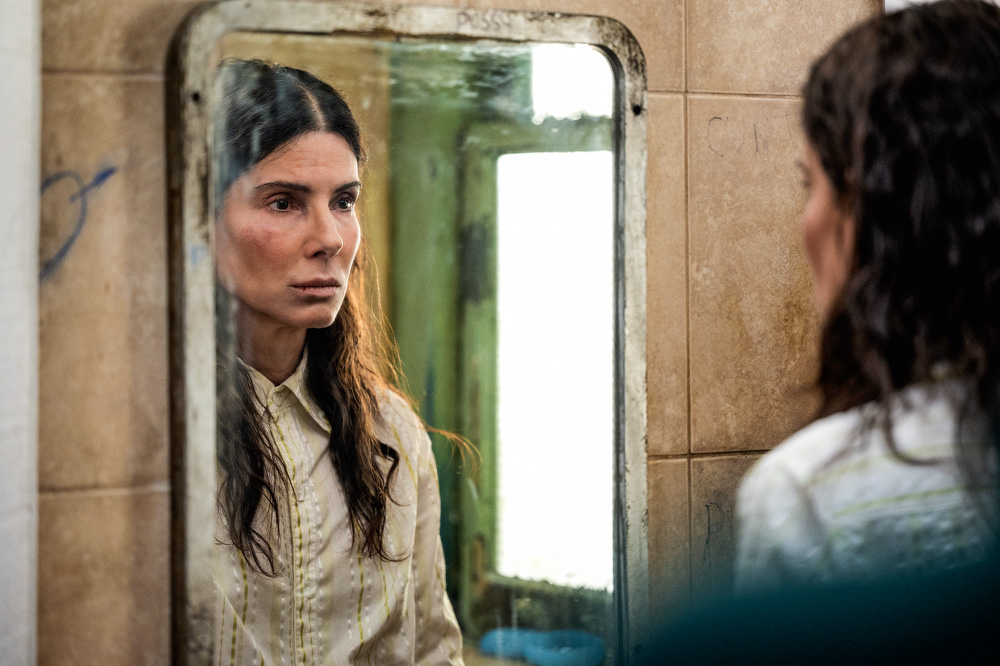 Imagem mostra mulher com camisa branca se olhando no espelho. O espelho é sujo e antigo como a parede em que ele está pendurado.