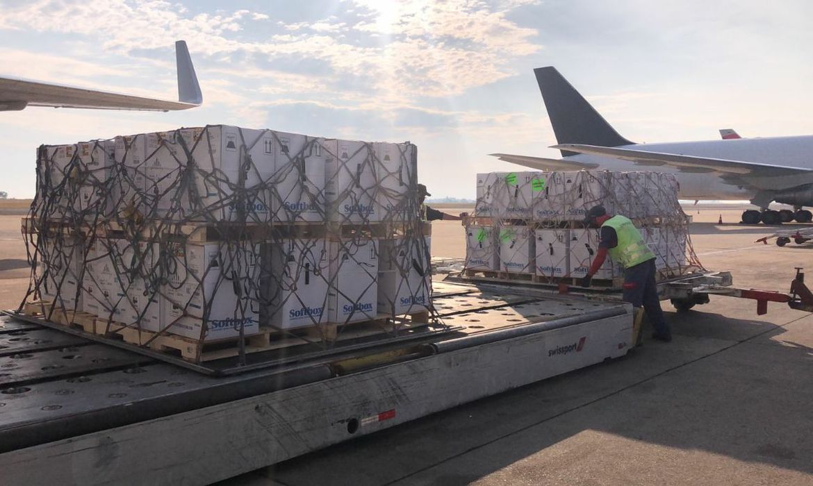 A foto mostra o carregamento da vacina que acabou de chegar no aeroporto. Um funcionário, que traja um uniforme verde fluorescente, mexe na carga.