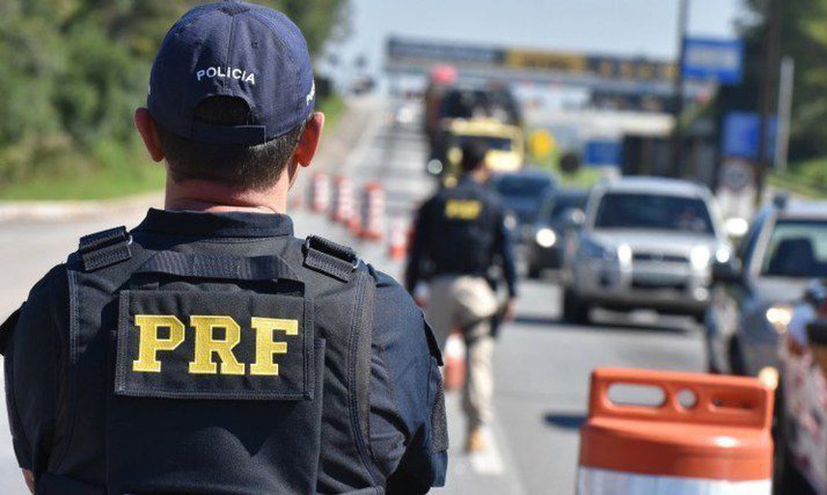 Imagem mostra dois policiais rodoviários federais durante uma blitz, com cones e carros