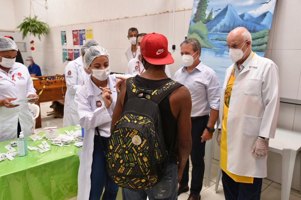 Na foto uma pessoa é vacinada contra a Covid-19. Ao lado da equipe de enfermagem estão o secretário municipal da Saúde Edson Aparecido e o Padre Júlio Lancellotti.