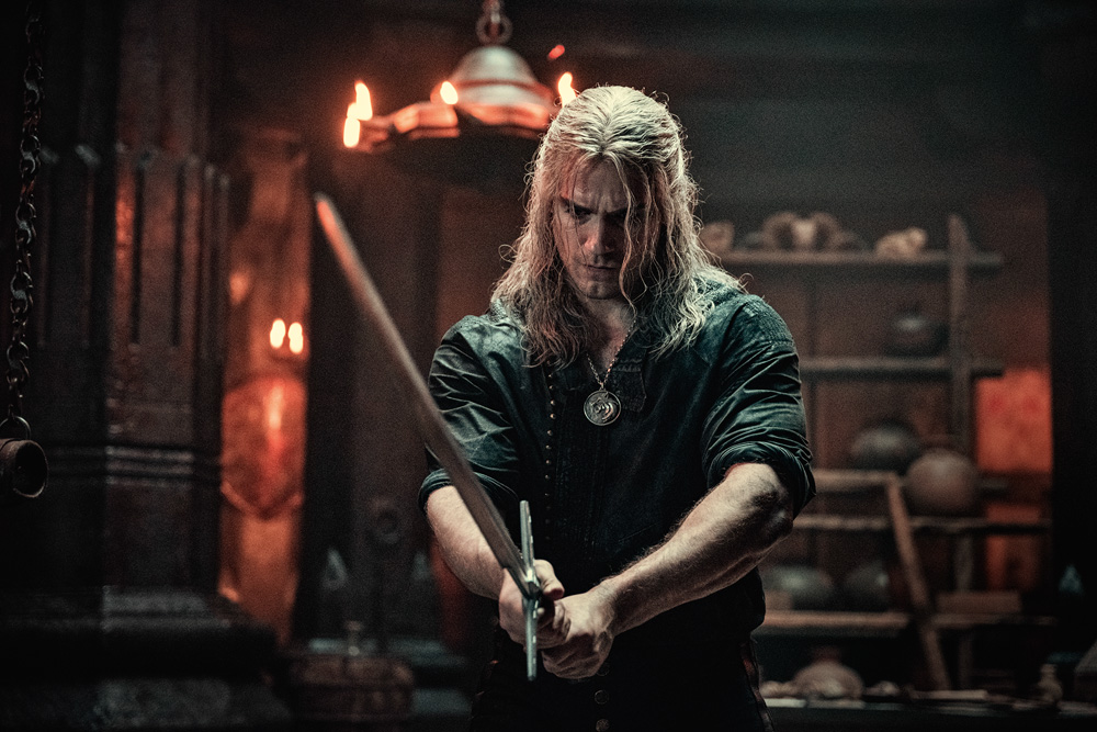 Imagem mostra homem de de cabelos brancos longos segurando uma espada em um cômodo escuro.