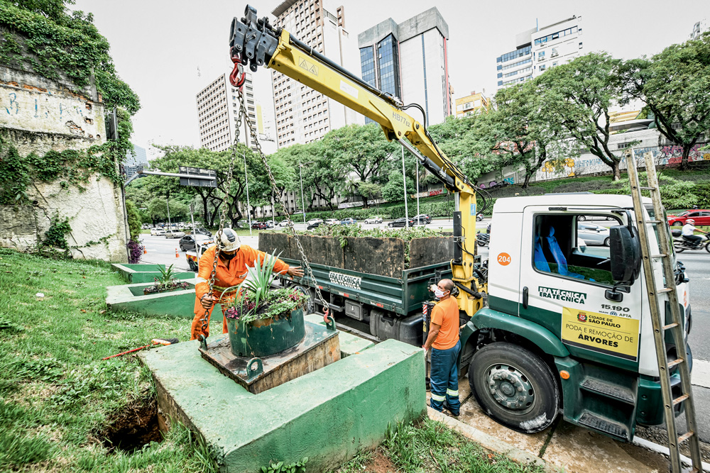 Imagem mostra guindaste levantando bloco de concreto com vaso de planta em cima. Dois homens vestindo coletes laranjas ajudam o processo.