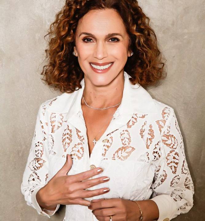 Retrato de Adri Coelho Silva, uma mulher de meia-idade, branca, de cabelos castanhos cacheados. Ela usa uma camisa rendada na cor branca.