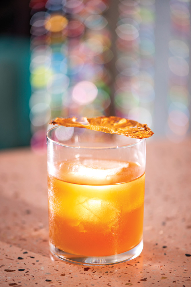 Drinque de cor alaranjada servido em copo baixo e de fundo redondo decorado por um pedaço de abacaxi desidratado