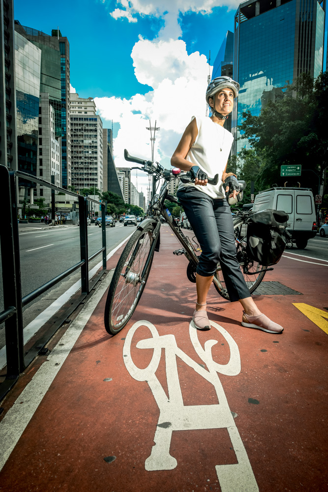 célia ciclovia avenida paulista Imagem mostra mulher de camiseta branca e calça preta apoiada sobre sua bicicleta, em uma ciclovia entre duas faixas de carro.