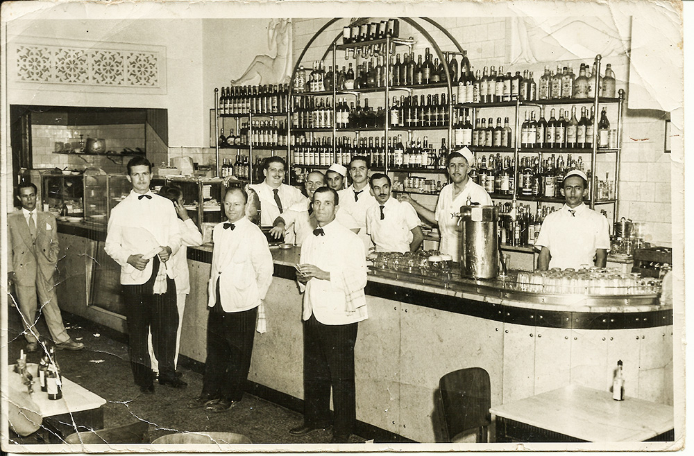 Equipe Década de 50 – Ponto Chic Imagem mostra balcão de bar com diversos garçons de camisas brancas, calças prestas e gravatas borboletas.