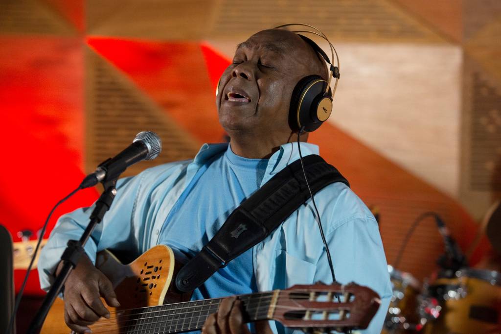 Filó Machado -foto de Eugenio Goulart Imagem mostra homem de camisa e camiseta azul segurando um violão e cantando em um microfone. Ele também usa um fone.