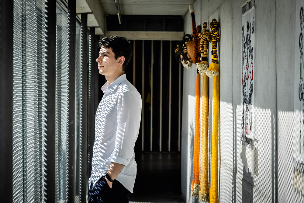 Franco Leme, um jovem branco e de cabelos escuros, posa em frente a uma janela da galeria. Ele usa uma camisa branca e raios de sol atingem seu rosto.