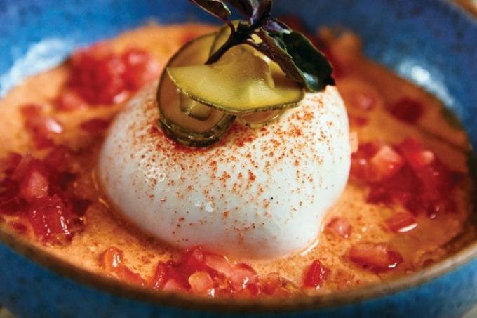 Sopa avermelhada com pequenos pedaços de tomate e servida junto de burrata com rodelas de abobrinha