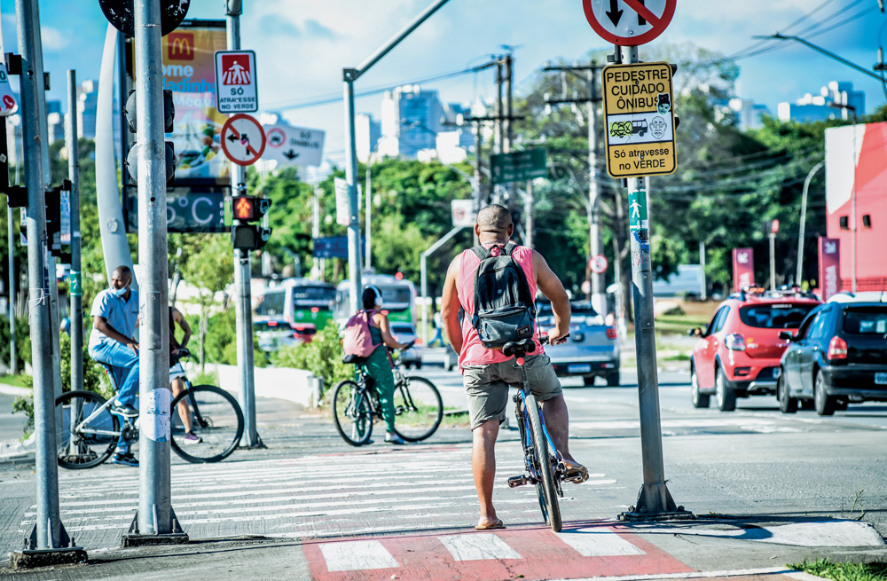 Imagem mostra três ciclistas, um em foco e os demais desfocados, esperando cada um de um lado da rua para atravessar na faixa. Ao fundo, postes, árvores e prédios.