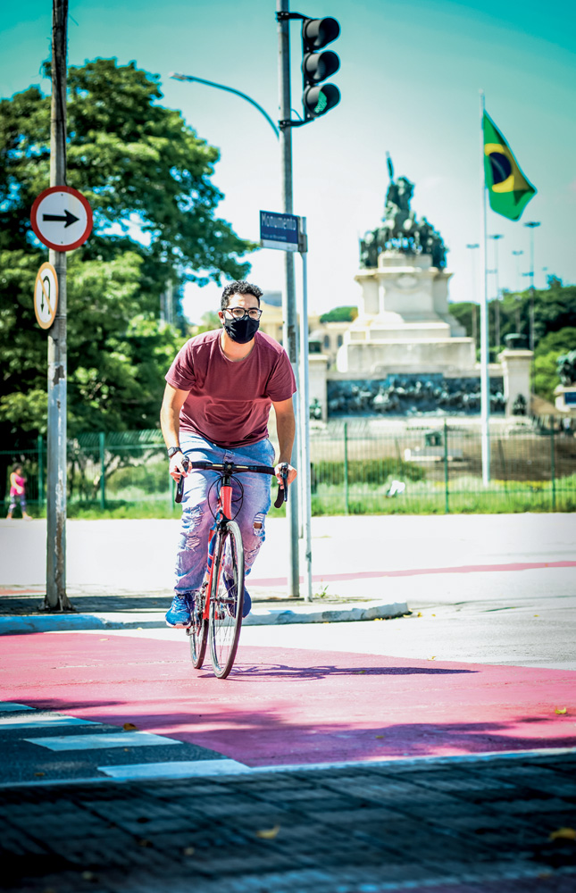 Imagem mostra homem de calça jeans e camiseta vinho atravessando a rua em uma ciclovia. Ao fundo, um monumento.