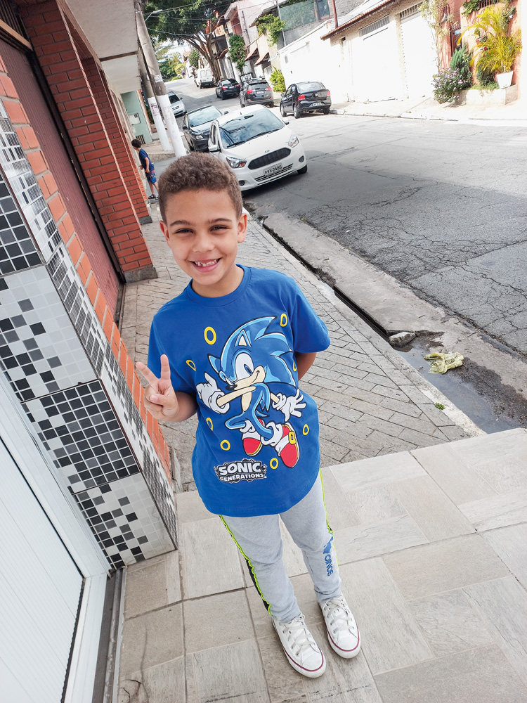 JUANfoto.jpg Juan aparece na rua e faz um sinal para a foto. Usa uma camiseta azul estampada e uma calça jeans.