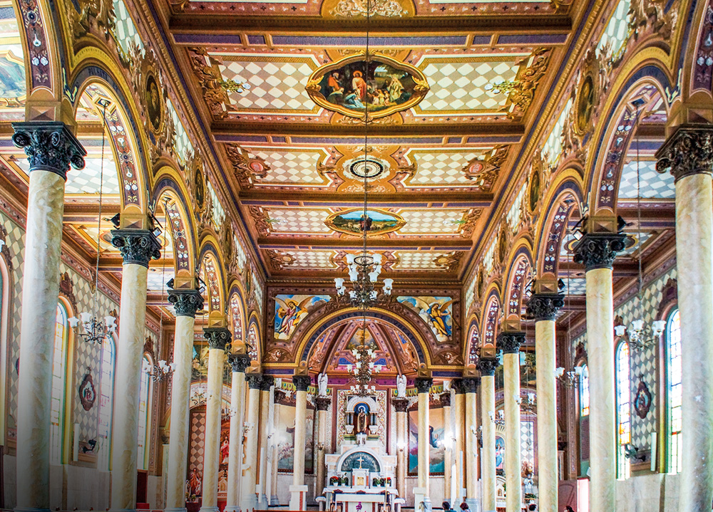 P257 Santa Teresinha – Percival Tirapeli.jpg Imagem mostra salão de igreja com teto marrom e branco, com imagens bíblicas entre padrões quadriculados. Duas fileiras de colunas de mármore acompanham o salão até o fundo, onde está um altar.