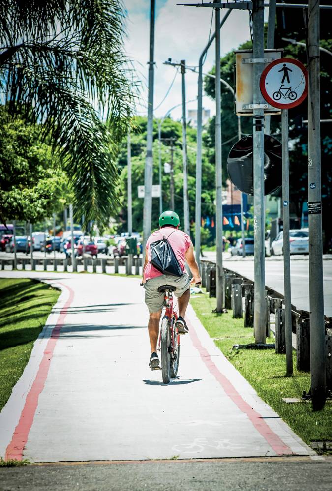 Imagem mostra ciclista de camiseta vermelha andando em ciclovia cinza, entre um gramado e uma avenida.