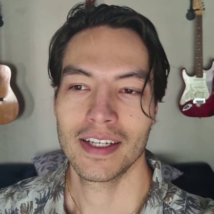 Imagem msotra homem de cabelo molhado com a boca entreaberta em frente a parede com um violão e uma guitarra pendurados.