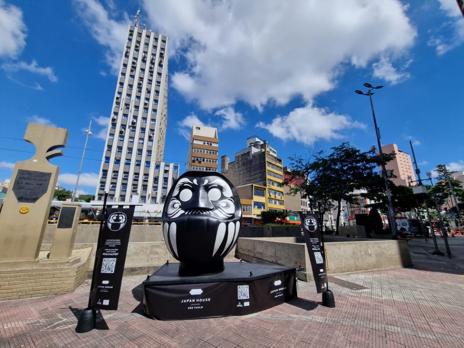 daruma Imagem mostra escultura preta com traços brancos formando um rosto. Ela está sobre uma plataforma e em uma praça com prédios ao fundo.
