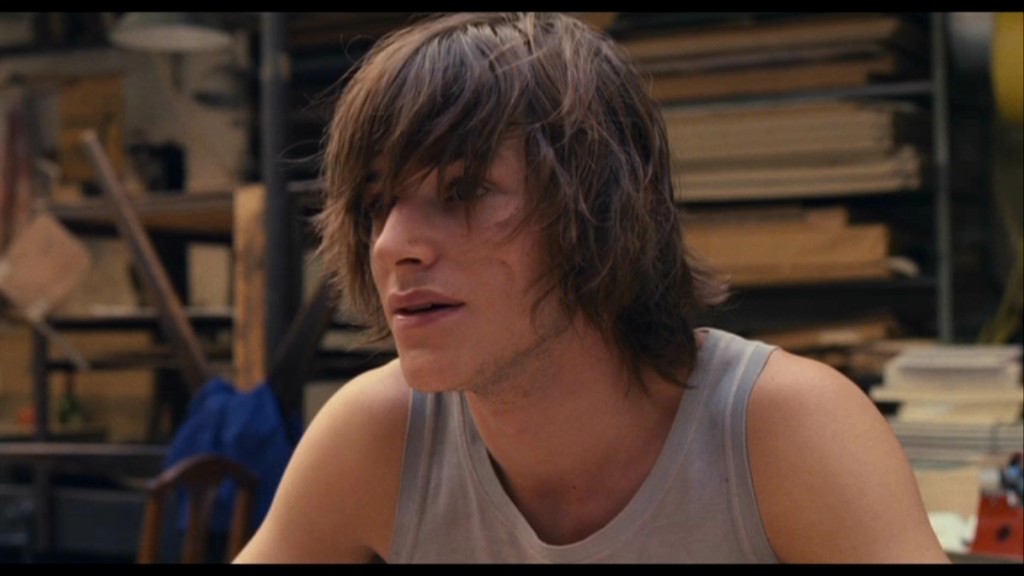 gaspard_ulliel_1290614255 Imagem mostra homem de cabelo comprido e com franja com regata cinza.