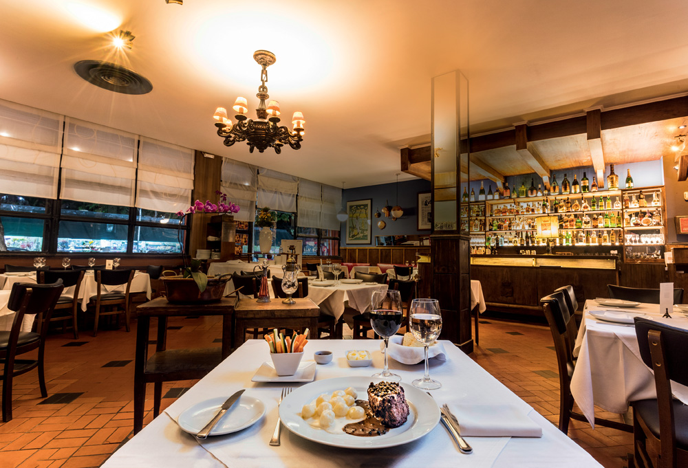 Foto Paulo Vitale Imagem mostra espaço com mesas e cadeiras, uma parede azul ao fundo e janelas. À frente na foto está um prato com carne e batatas e duas taças, uma de vinho e outra com água.