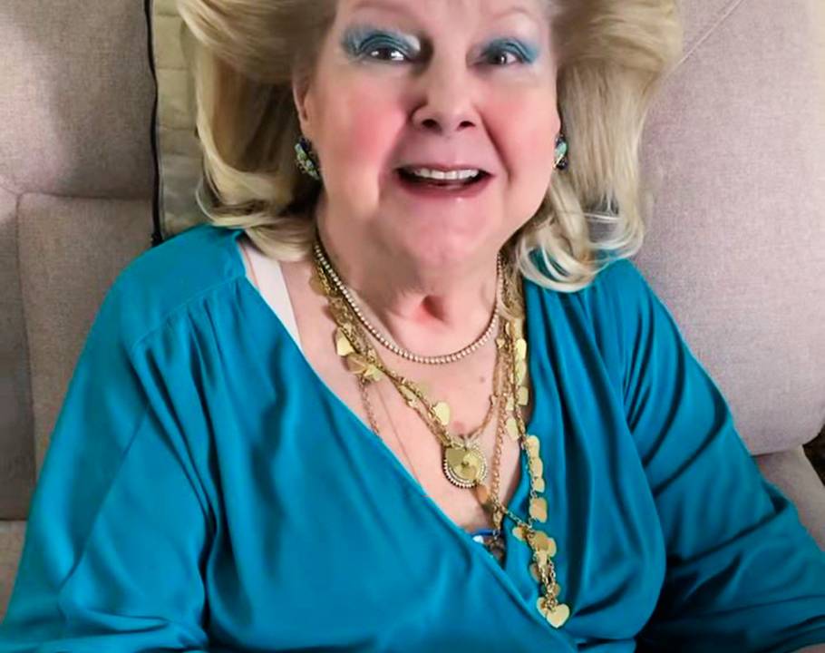 Foto mostra dona Maricy , loira, de maquiagem azul, usando uma blusa também azul. No pescoço, muitos colares dourados.
