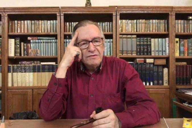 Foto mostra Olavo de Carvalho, um senhor branco, de óculos e cabelos grisalhos, sentado. Ao fundo, há uma prateleira cheia de livros.