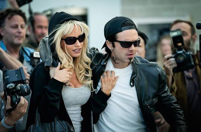 pam e tommy – credito divulgacao.JPG Imagem mostra uma mulher e um homem, ambos de óculos escuros e jaqueta de couro, abraçados, caminhando entre fotógrafos.