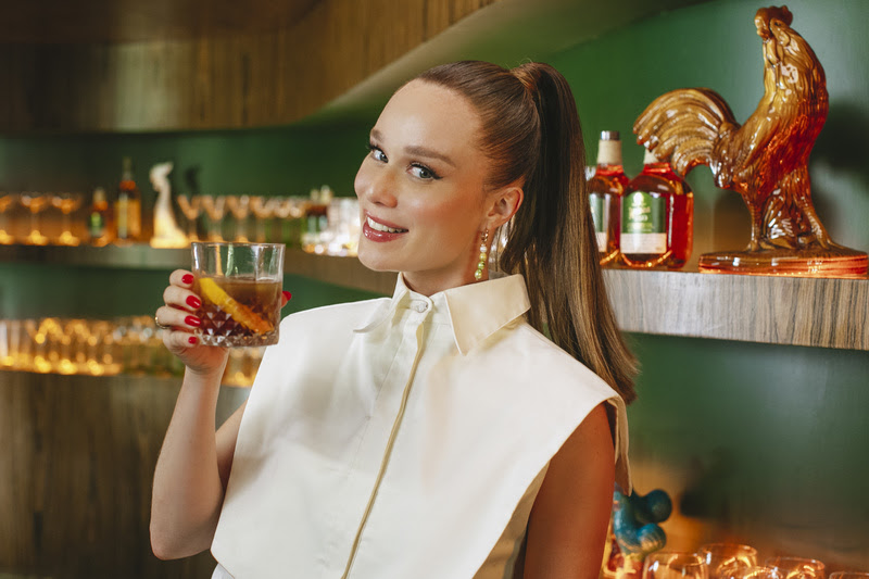 Mulher loira usa vestido branco e cabelo preso enquanto pose segurando um copo com um drinque de cor amadeirada