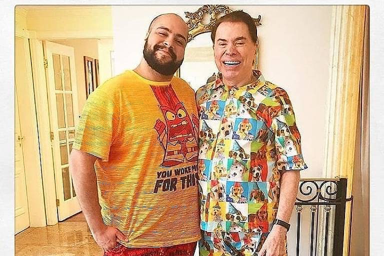 Imagem mostra dois homens sorrindo, em pé, um ao lado do outro. O da esquerda veste uma camiseta amarela e vermelha, e o da direita veste uma camisa colorida e estampada.