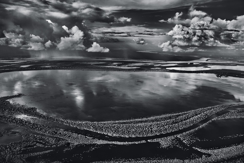 Imagem em preto e branco mostra paisagem com diversos corpos d'água próximos. No céu, nuvens até o horizonte.