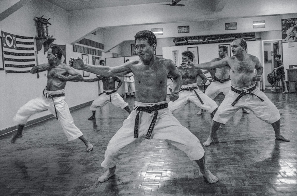 Foto em preto e branco mostra o sensei Taketo Okuda ensinando caratê para seus alunos. Todos estão sem camisa, com trajes tradicionais da modalidade. Na parede da academia, vê-se uma bandeira de São Paulo.