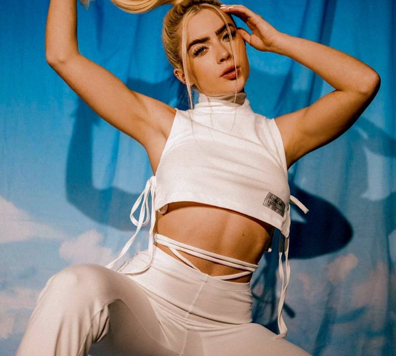 Jade Picon aparece agachada. Ela veste um conjunto branco com cropped e calça comprida. Ela é uma mulher branca, loira e usa um rabo de cavalo. O fundo do cenário é um céu azul com nuvens.