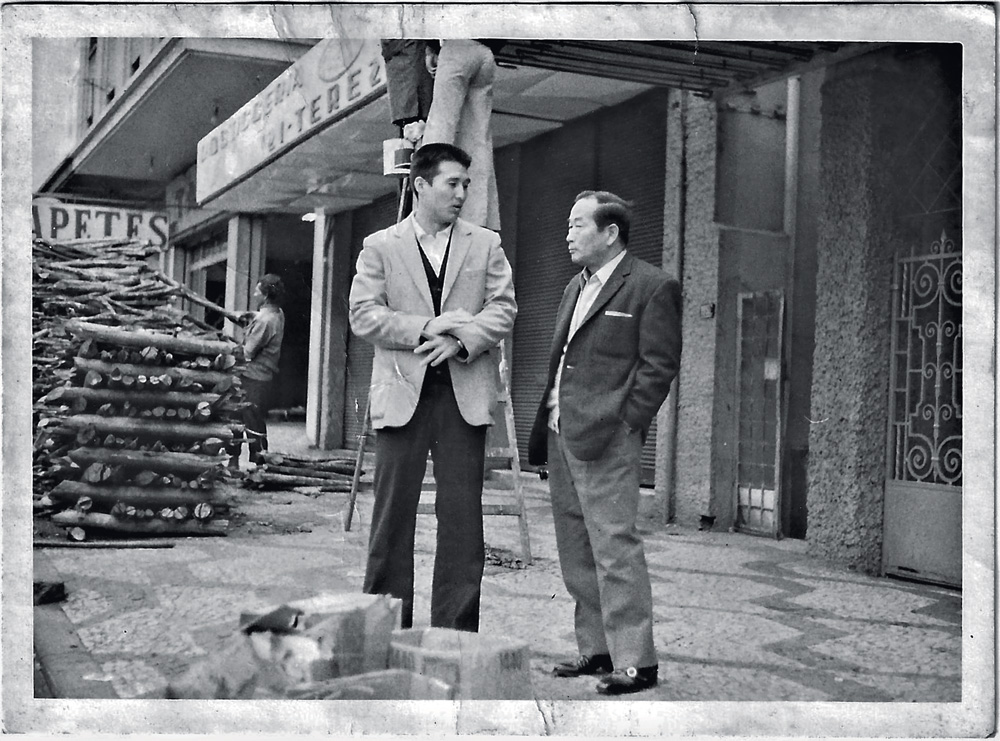 4.jpg Foto em preto e branco mostra dois homens japoneses nas ruas de São Paulo. Ambos trajam um terno e conversam entre si.