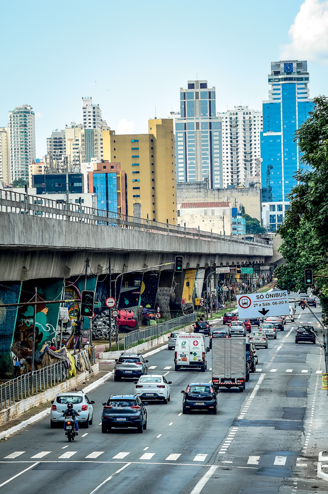 Imagem mostra avenida cheia de carros. Ao lado, um viaduto e um horizonte de prédios ao fundo.