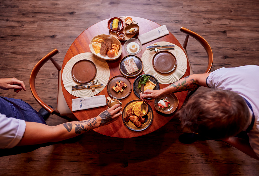 Mesa de madeira com diversas guarnições dispostas em seu topo. Também é possível ver duas pessoas se servindo