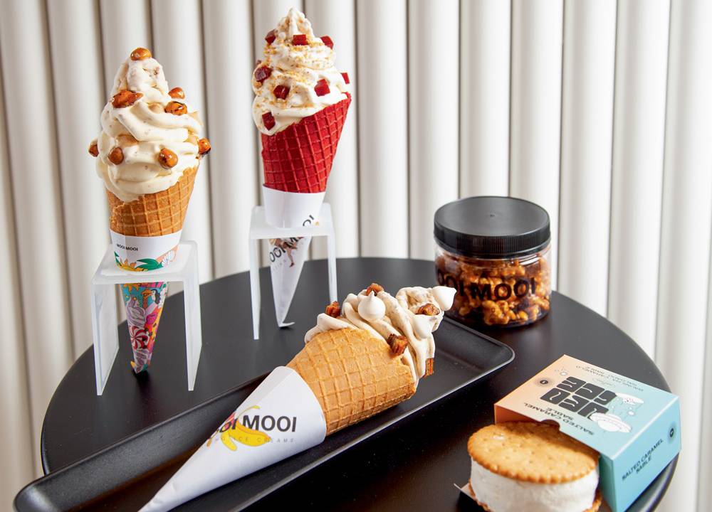 Três diferentes tipos de casquinha de sorvetes, sendo inclusive uma delas de cor vermelha, um biscoito recheado por sorvete e um pequeno recipiente de plástico sobre uma mesa de cor preta