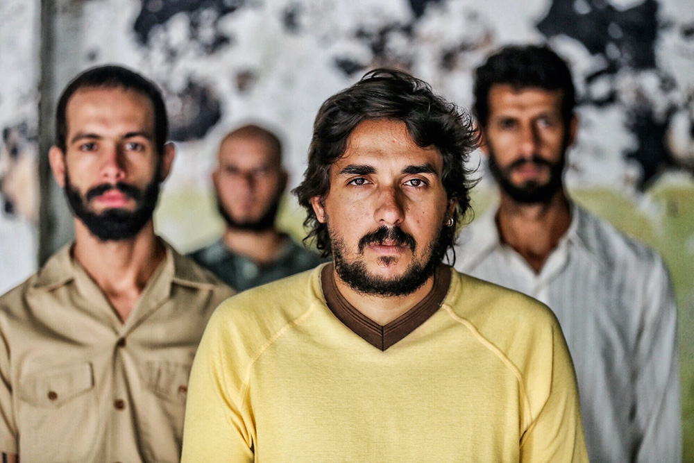 Imagem mostra quatro homens. Um está no foco da imagem, à frente, enquanto os demais estão ao fundo, desfocados.
