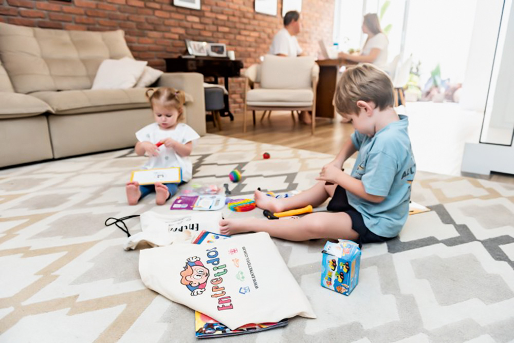 Foto mostra duas crianças pequenas, uma menina e um menino, sentados no chão brincando com várias atividades espalhadas pelo chão. Ao fundo, pai e mãe estão sentados em uma mesa.