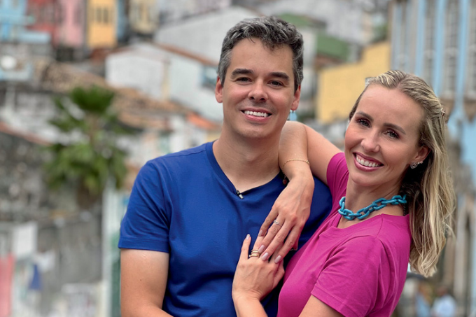 Imagem mostra um homem de camiseta azul e uma mulher de roupa rosa abraçados, sorrindo para a câmera.