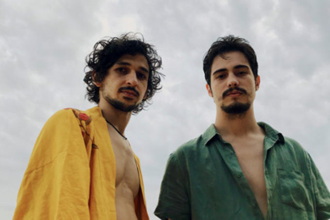 Imagem mostra dois homens, um de camisa amarela e o outro de camisa verde. Eles olham para a câmera.