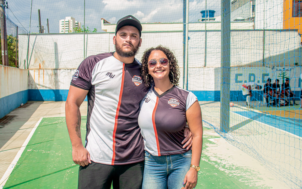 Imagem mostra homem e mulher abraçados. Eles vestem uma camiseta igual, metade branca e metade preta. Eles estão em uma quadra de futsal. É possível ver alguns jogadores sentados em um banco, à direita.