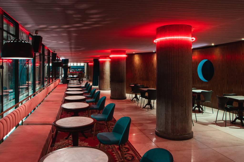 Segundo andar do Riviera Bar decorado por mesas de mármore branco, poltronas de estofado azulado e luzes vermelhas de LED