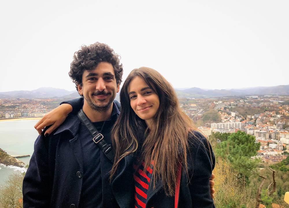 Foto de Camila e o noivo. Ambos são brancos, de cabelos e olhos escuros. Ela tem o cabelo liso e comprido e ele o cabelo enrolado e barba. Eles posam abraçados em frente a uma paisagem. Ambos usam roupa de frio em tons escuros.