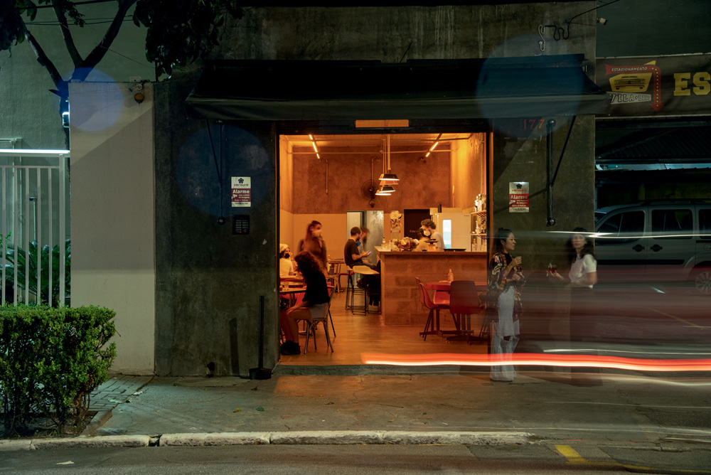 Fachada de um bar. O interior do ambiente está iluminado com uma luz aconchegante. Algumas pessoas circulam lá dentro.