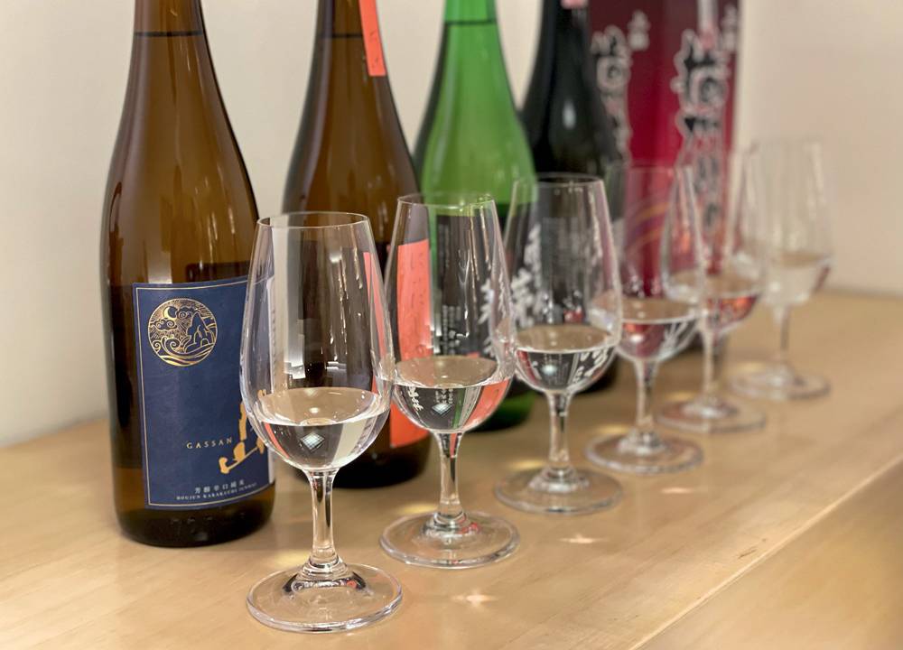 Garrafas de saquê alinhadas sobre mesa de madeira com taças de cristal com a bebida servida na frente de cada uma.