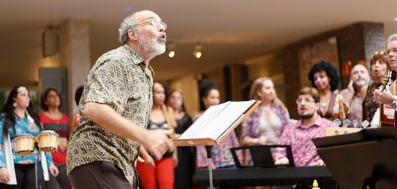 Imagem mostra homem grisalho de camisa com partitura em sua frente. Ao fundo, um grupo de pessoas cantando e tocando instrumentos.