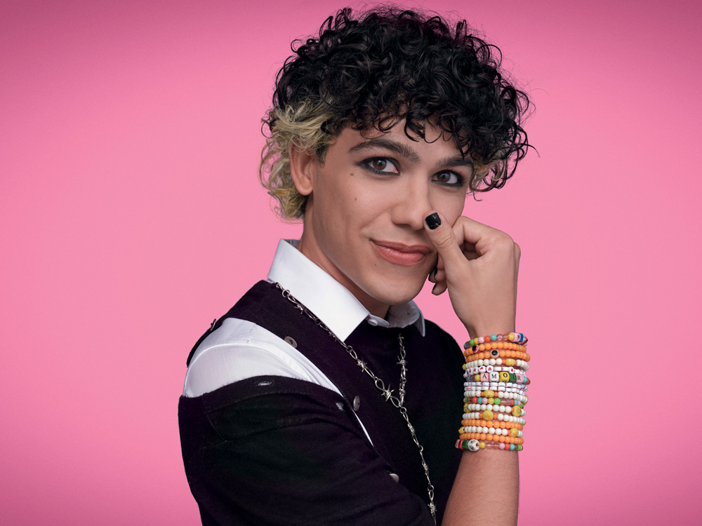 de volta aos 15 – credito Gustavo Zylbersztajn (1).jpg Imagem mostra pessoa com cabelo preto com mechas descoloridas atrás. Ela veste uma roupa preta e posa em frente a um fundo rosa.