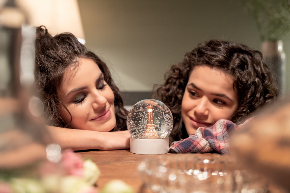 Imagem mostra duas meninas debruçadas sobre mesa de madeira, olhando para um globo de neve, entre elas.