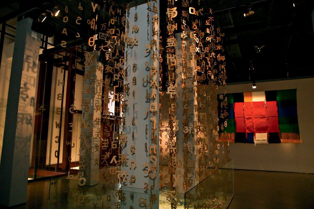 Imagem mostra exposição com diversas letras douradas de alfabetos diferentes penduradas por fios.