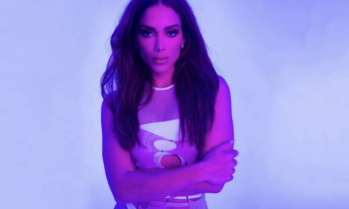 Anitta é uma mulher branca, de cabelos lisos e escuros. Usa um body branco e está sob uma luz roxa em seu clipe