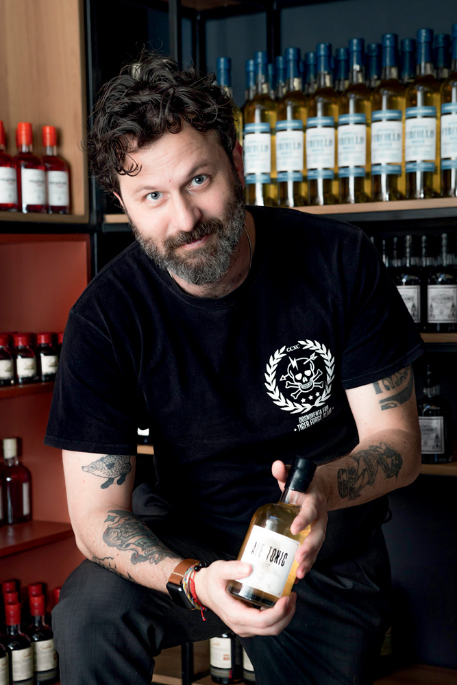 Raiz Club by APTK Spirits Alê D’Agostino posa segurando uma garrafa de um drinque engarrafado